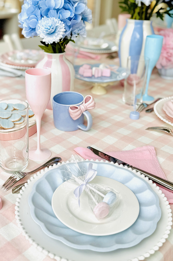 Table dressée en pastel rose et bleu : assiettes superposées sur une nappe à carreaux, serviette rose, hortensias bleus et faveurs emballées - Table by At Home With Soni