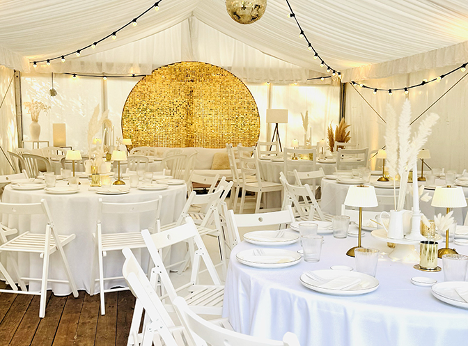 Chapiteau dressé pour une fête, tables rondes en blanc, chaises blanches, plumes de pampa, fond doré et guirlandes lumineuses - Table by At Home With Soni